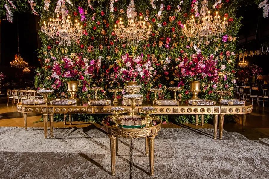 Mesa de doces dourada diante de painel de flores e lustres de cristal