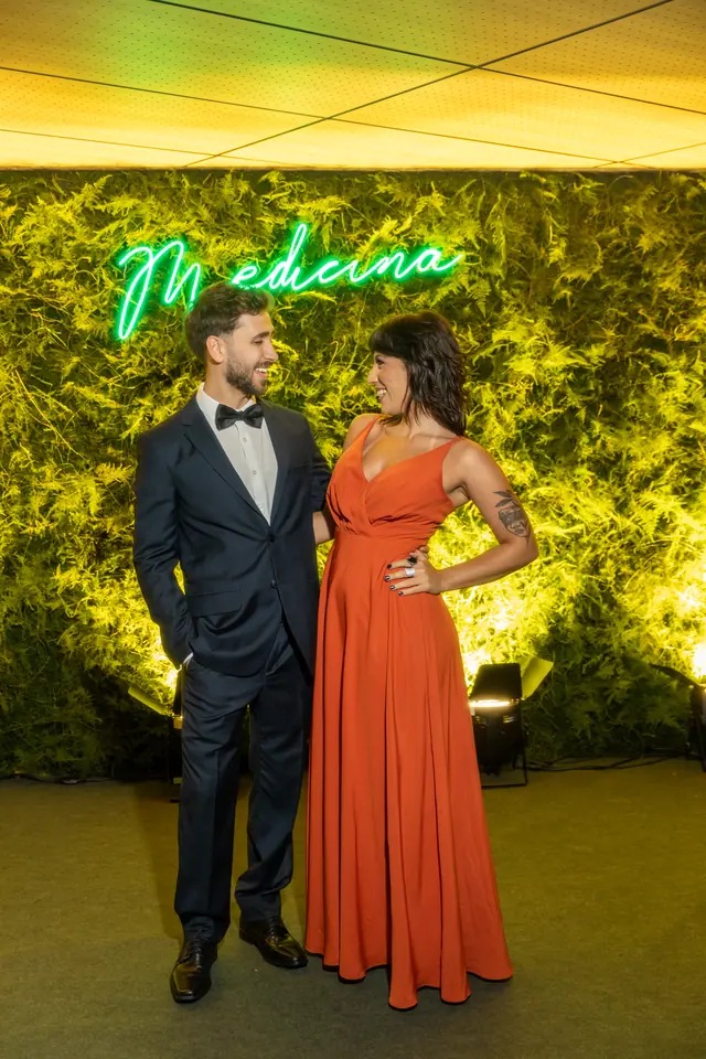 Casal de formandos em traje de gala diante de painel verde